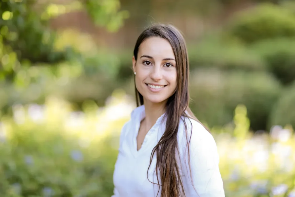 Portrait of Efrossini, a children’s book author, smiling outdoors with long brown hair, wearing a white blouse, set against a blurred green garden background.