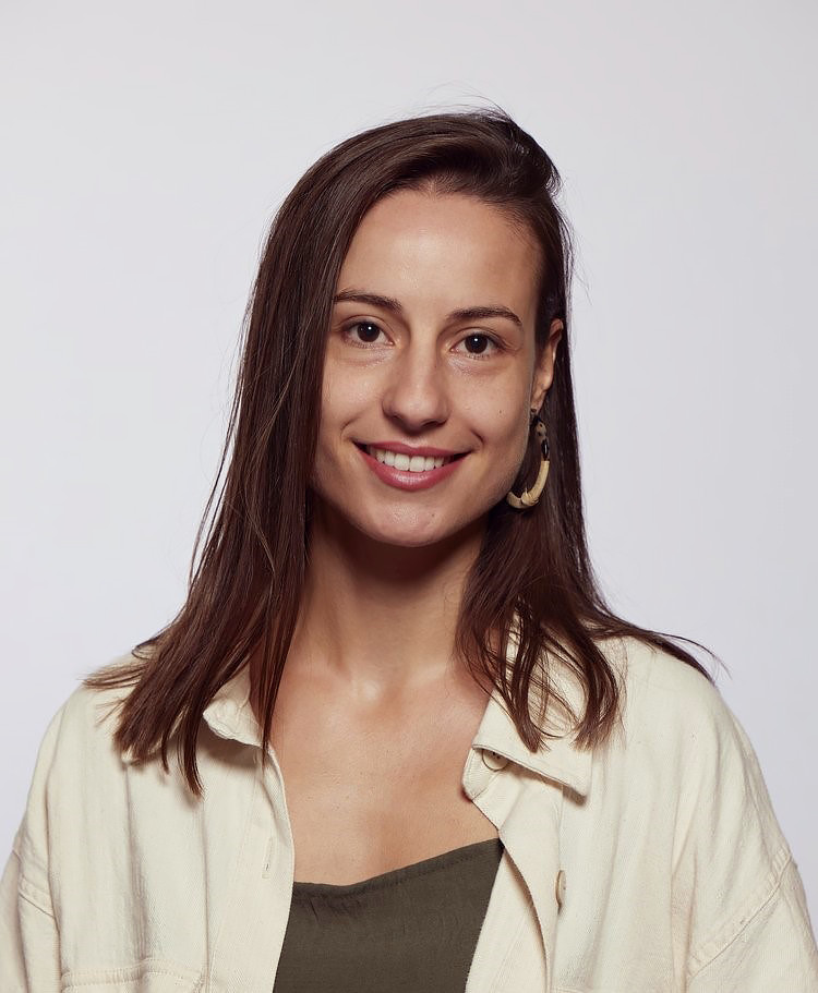 Portrait of Efrossini, a smiling children’s book author with long brown hair, wearing a cream-colored jacket over a dark top and large hoop earrings, against a plain white background.